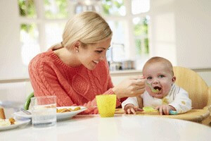 Young woman with blonde hair spoon feeding a baby at a table.