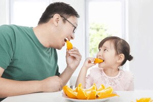 Male with dark hair and glasses eating orange with a young girl with dark hair.