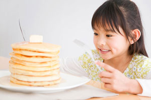 Young girl with dark hair holding a fork and knife looking at a large stack of pancakes.