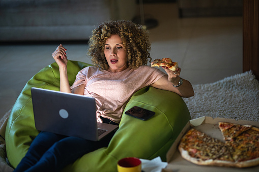 Woman with curly hair eating pizza, watching computer