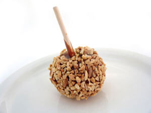 A caramel apple on a stick with nuts on it sitting on a white plate.