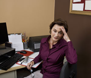 Woman in dark purple top looking away from her computer with her hand on her head.